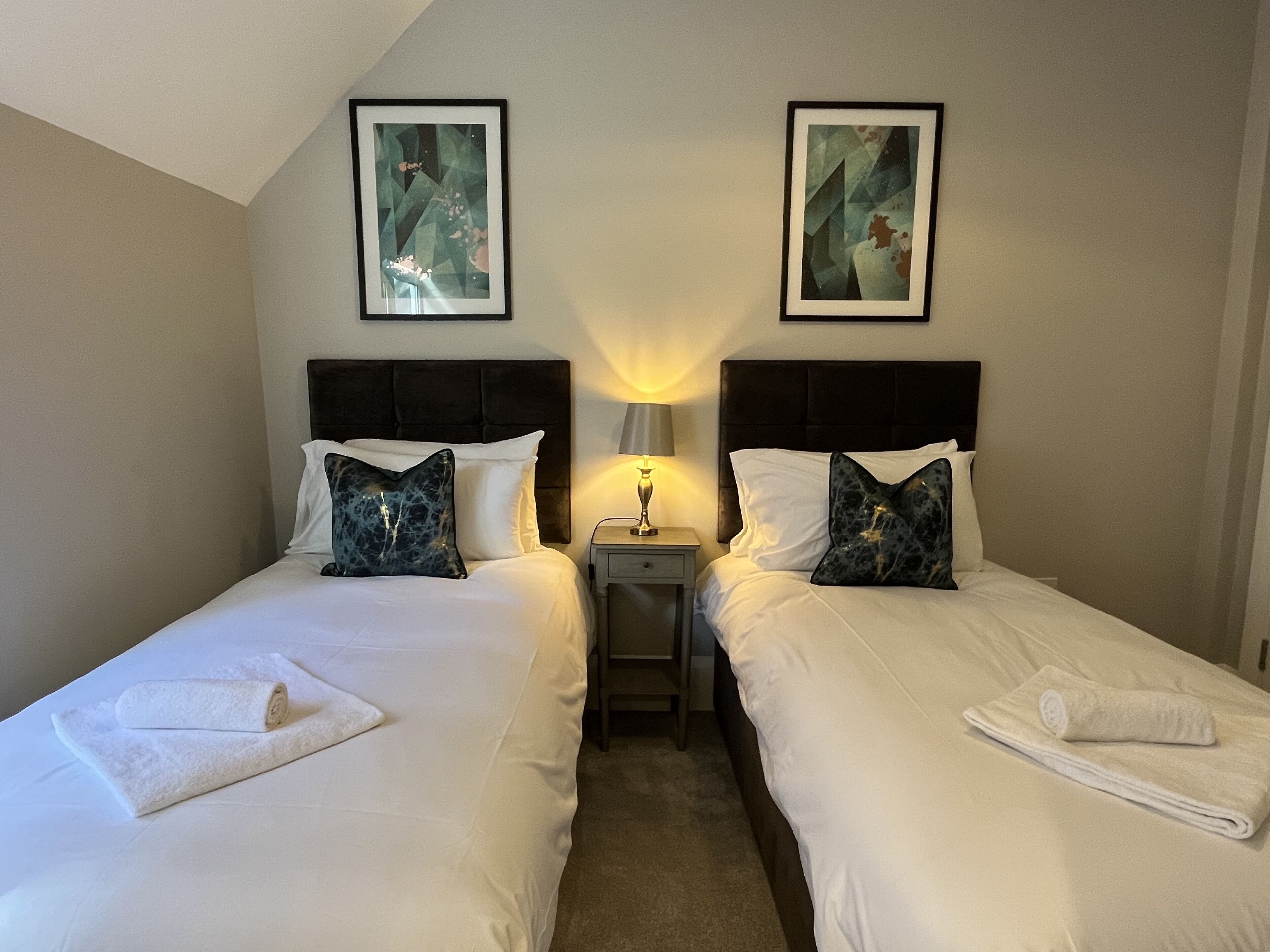 A hotel room with two beds, each with white linens and decorative pillows. There are framed artworks on the walls above the beds, and a small bedside table with a lamp between the beds.