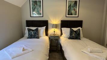 A hotel room with two beds, each with white linens and decorative pillows. There are framed artworks on the walls above the beds, and a small bedside table with a lamp between the beds.