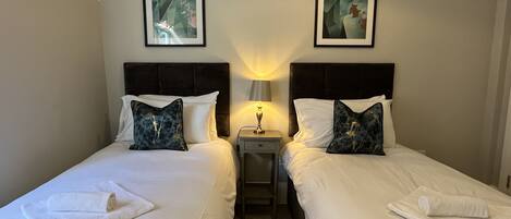 Een hotelkamer met twee bedden, elk voorzien van witte lakens en decoratieve kussens. Boven de bedden hangen kunstwerken in lijsten, en daartussen staat een klein nachtkastje met een lamp.