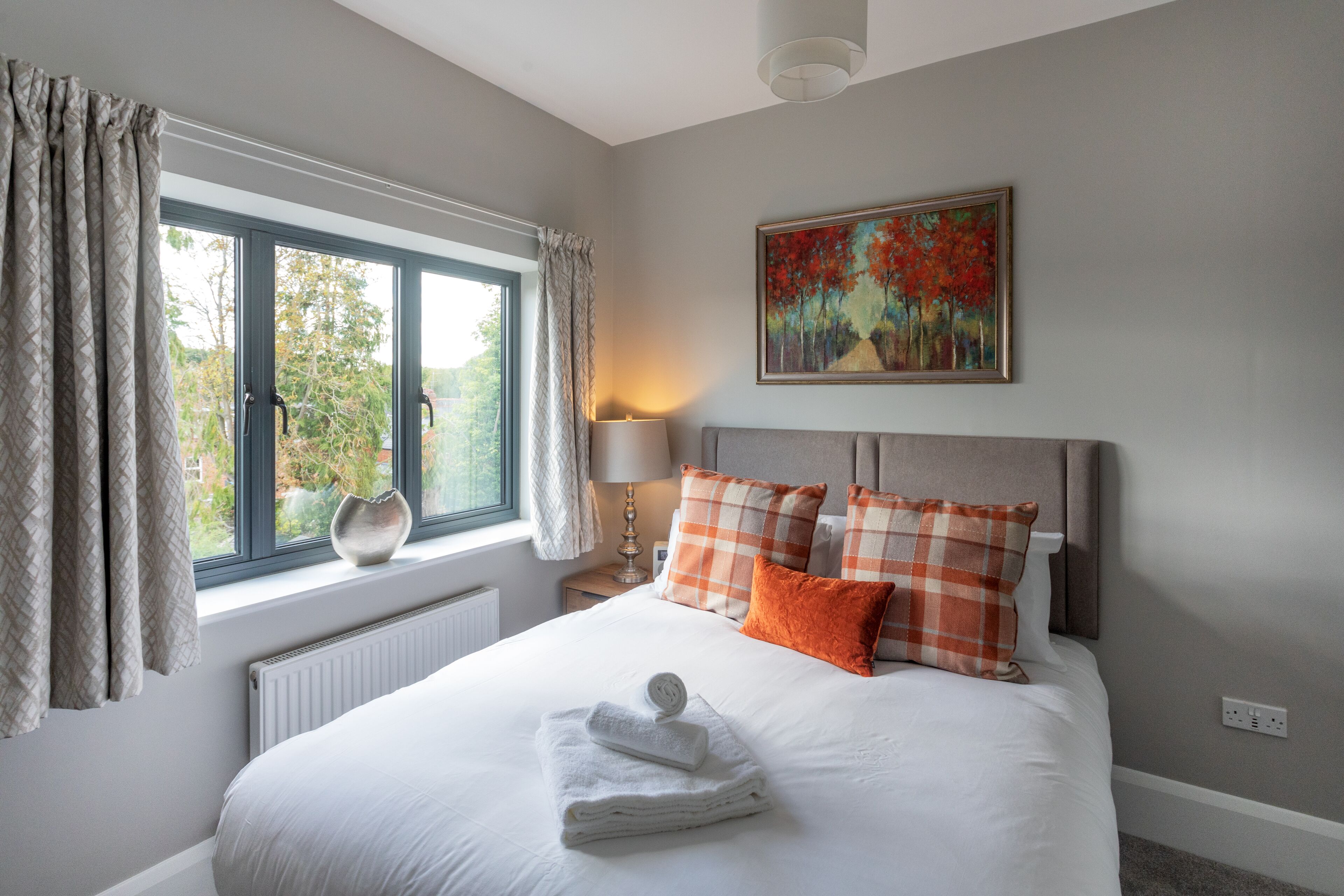 A neatly made bed with white linens and orange pillows, a window with curtains, a painting on the wall, and a bedside lamp.