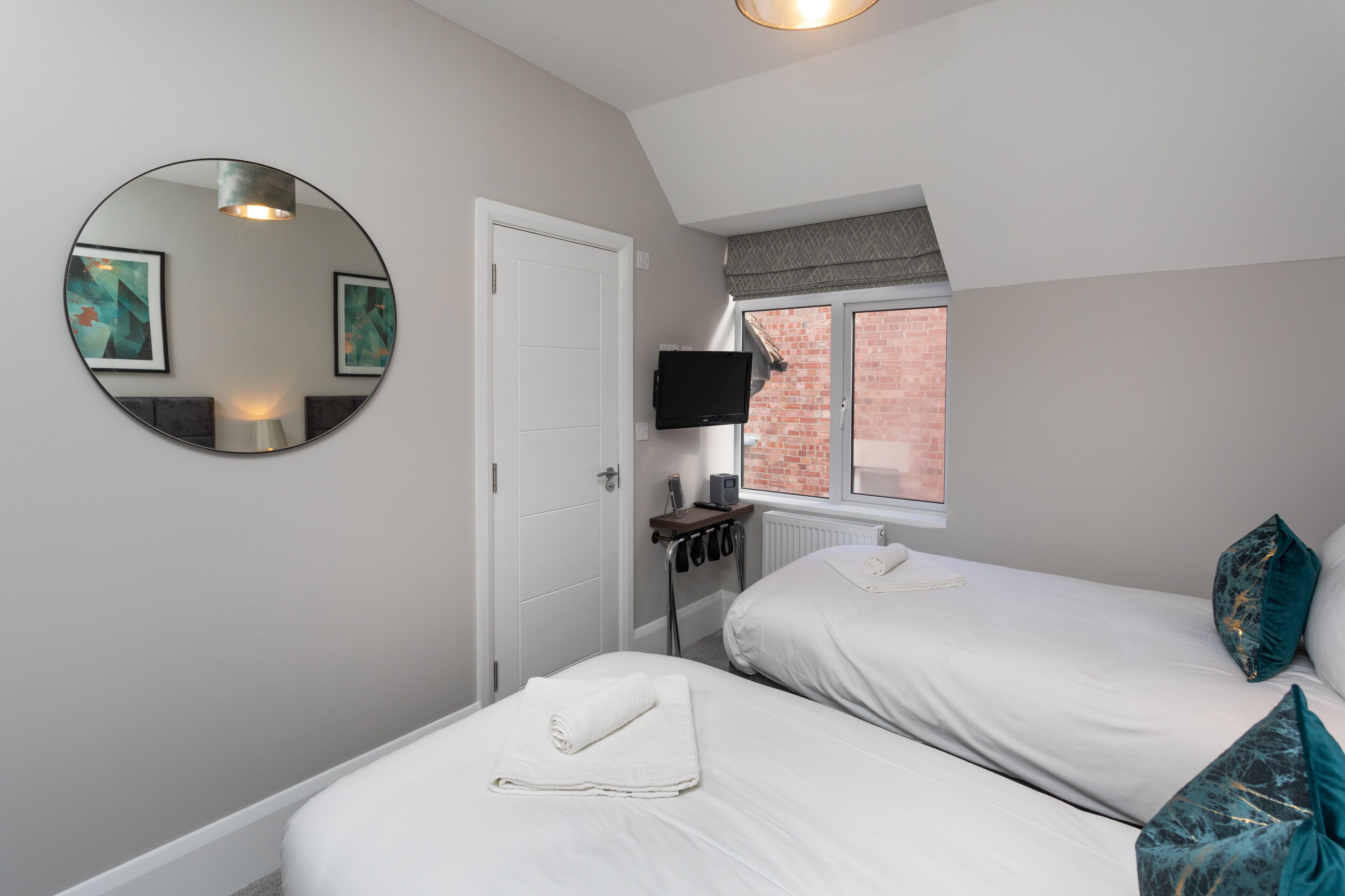 A bedroom with two beds, a round mirror, a television, and a window with a view of a brick building.