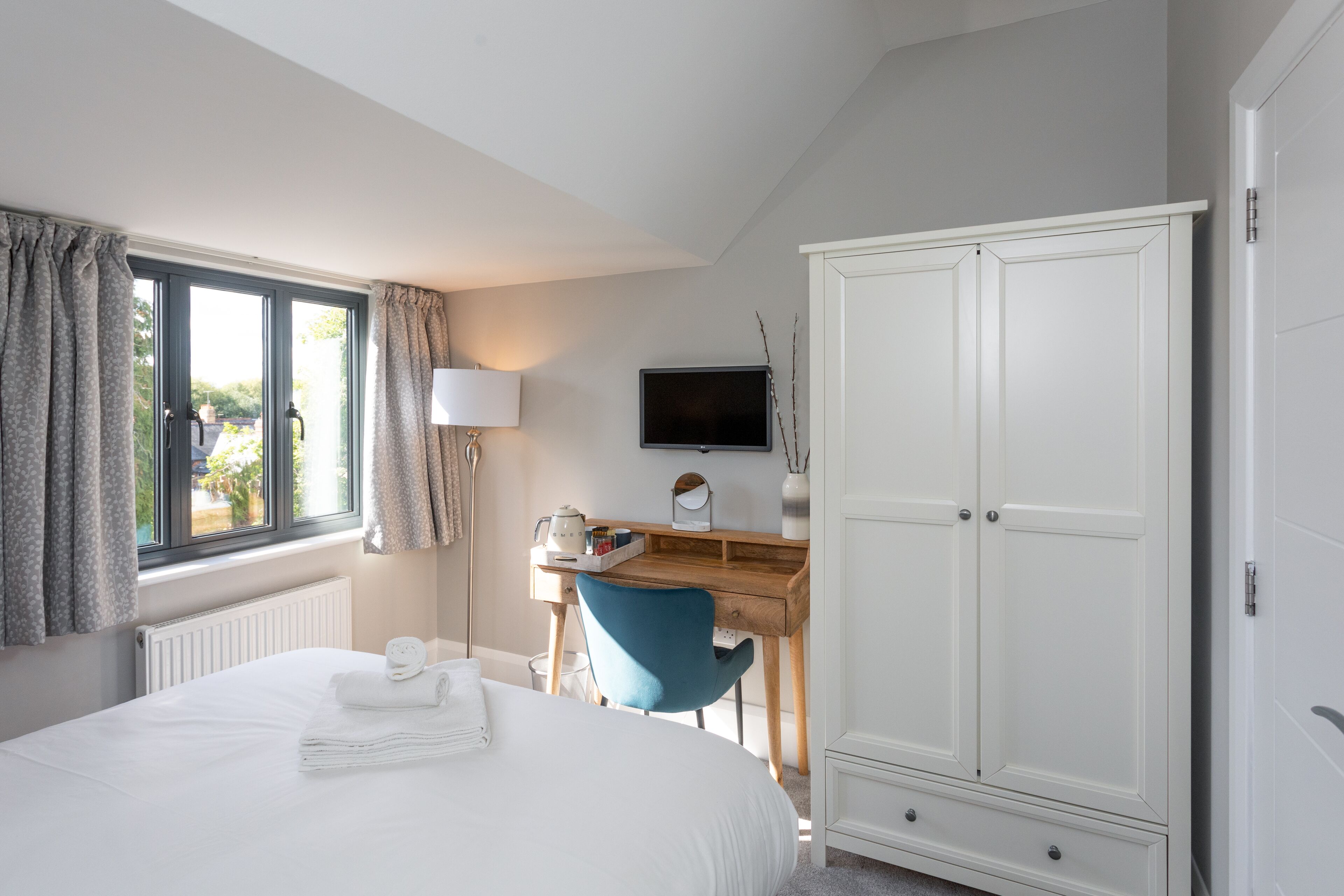 A bedroom with a bed, a desk with a chair, a wardrobe, and a television on the wall.