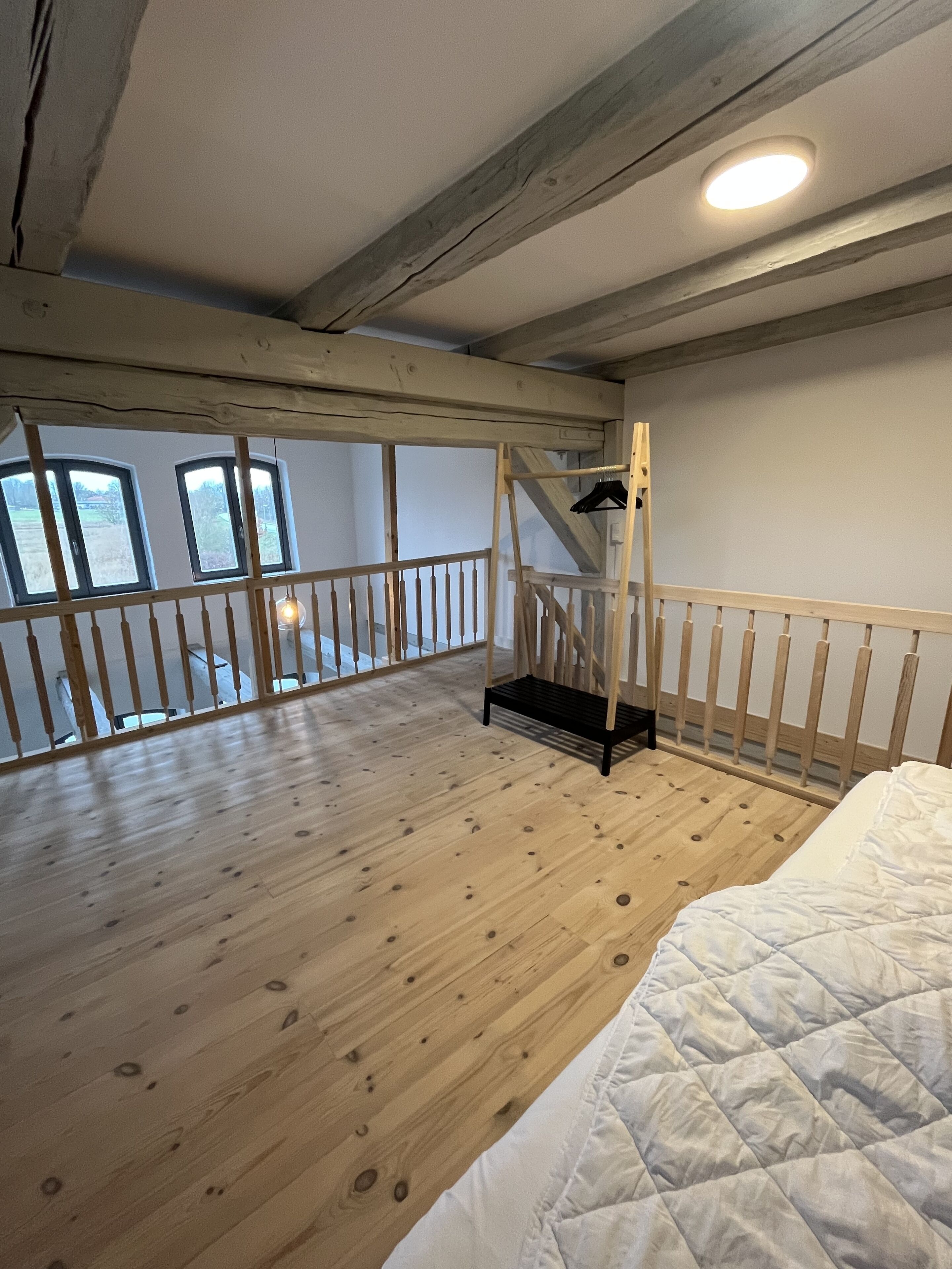 A spacious room with wooden flooring, a bed with a white duvet, a wooden railing, and a staircase leading to an upper level.