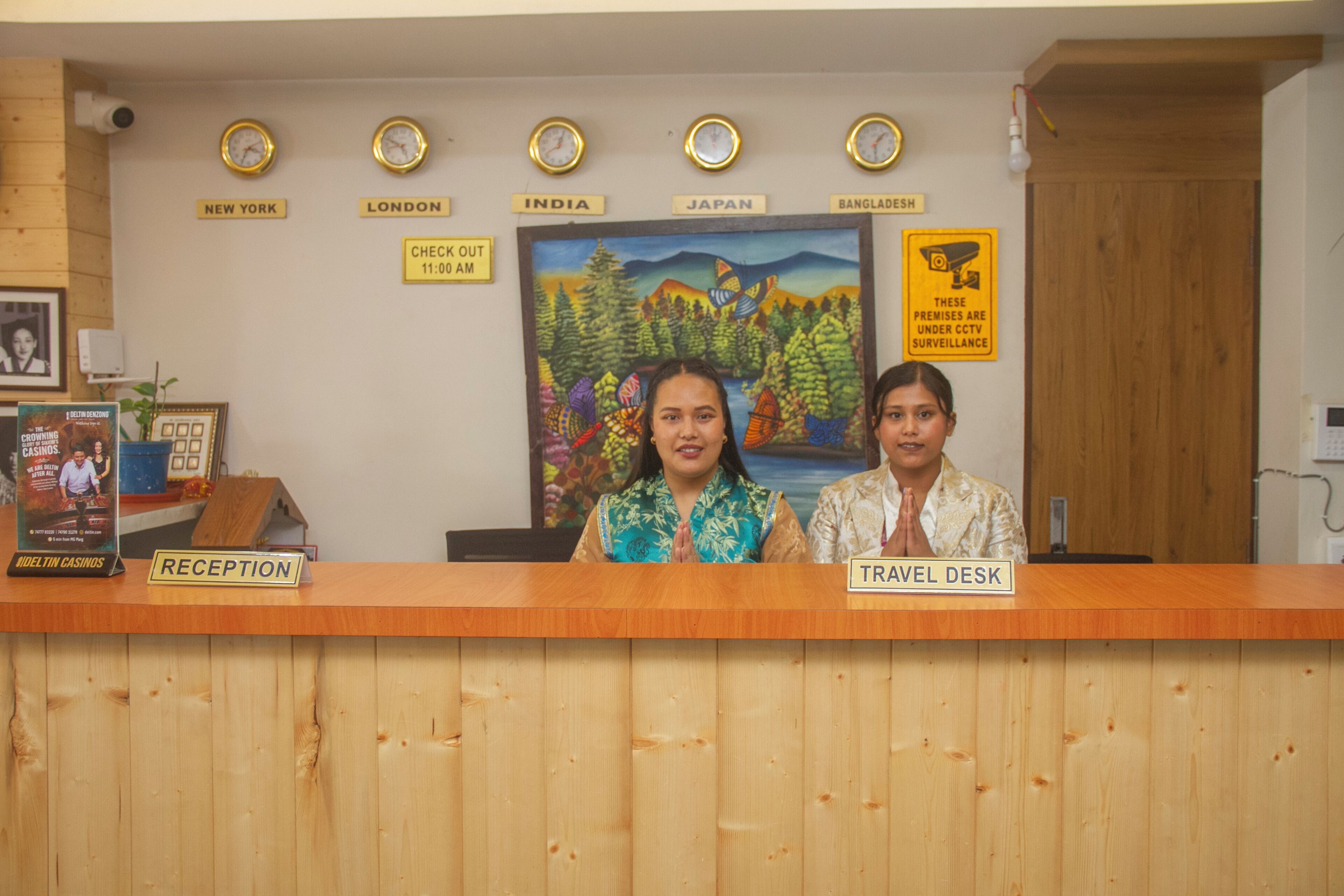 A reception desk with clocks displaying times for New York, London, India, Japan, and Bangladesh. There is a sign indicating check-out time and a CCTV surveillance warning.