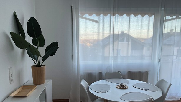 Een eethoek met een ronde tafel, witte stoelen, een potplant en uitzicht door lichte gordijnen.