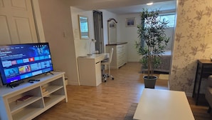 A living room with a TV on a stand, a white table, a potted plant, and a desk with a chair.