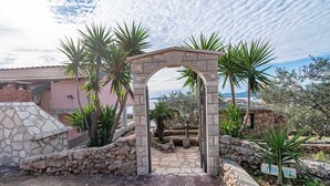A stone archway leading to a courtyard with a stone floor, surrounded by palm trees and a stone wall. - Villa Sea Edge (Orebic)