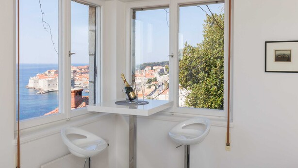 A corner room with a view of the sea and buildings, a small table with a bottle and glasses, and two white stools. - Apartment Rea (Dubrovnik)