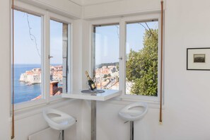 A corner room with a view of the sea and buildings, a small table with a bottle and glasses, and two white stools.