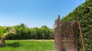 Une cabine de douche extérieure en bois, surmontée d’un toit en métal, entourée d’une terrasse dallée de pierres et d’une végétation luxuriante.