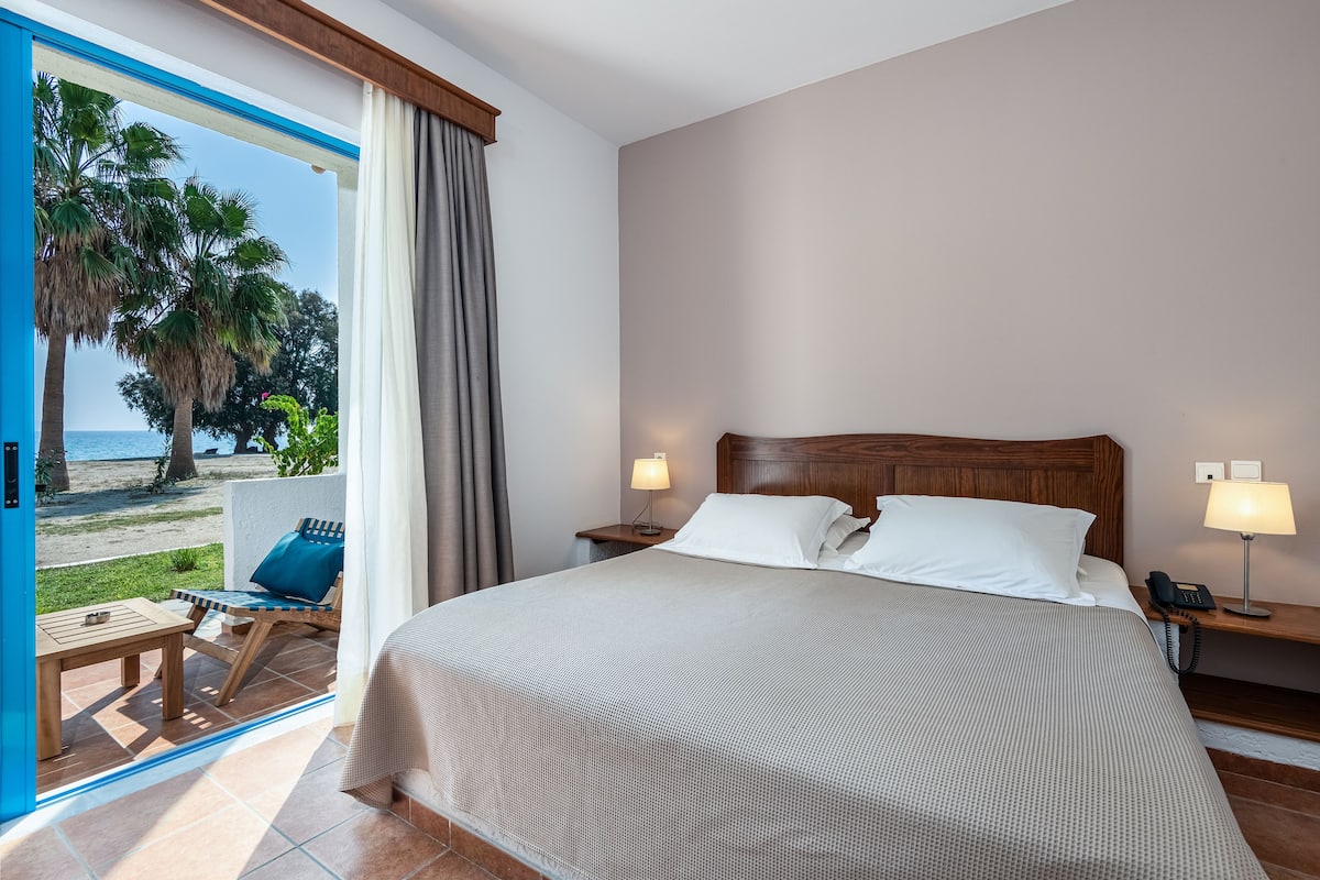 A hotel room with a large bed, two bedside tables with lamps, a wooden nightstand, a view of the beach, and a patio area with a table and chairs.