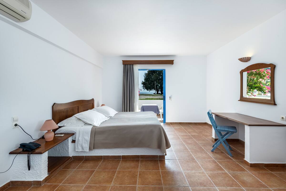 A bedroom with a bed, bedside table, lamp, desk, chair, and a view of a tree through a window.