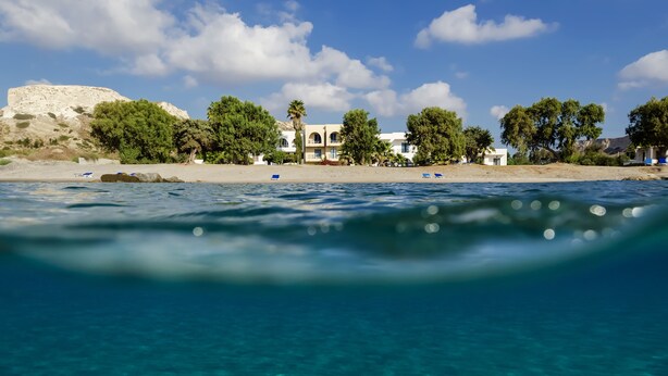 A beach view with clear water, a white building with a balcony, and trees. - Olympia Mare (Kos)