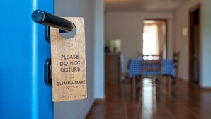 A wooden sign on a door with a message to not disturb, and a view of a dining area with a table and chairs. - Olympia Mare (Kos)