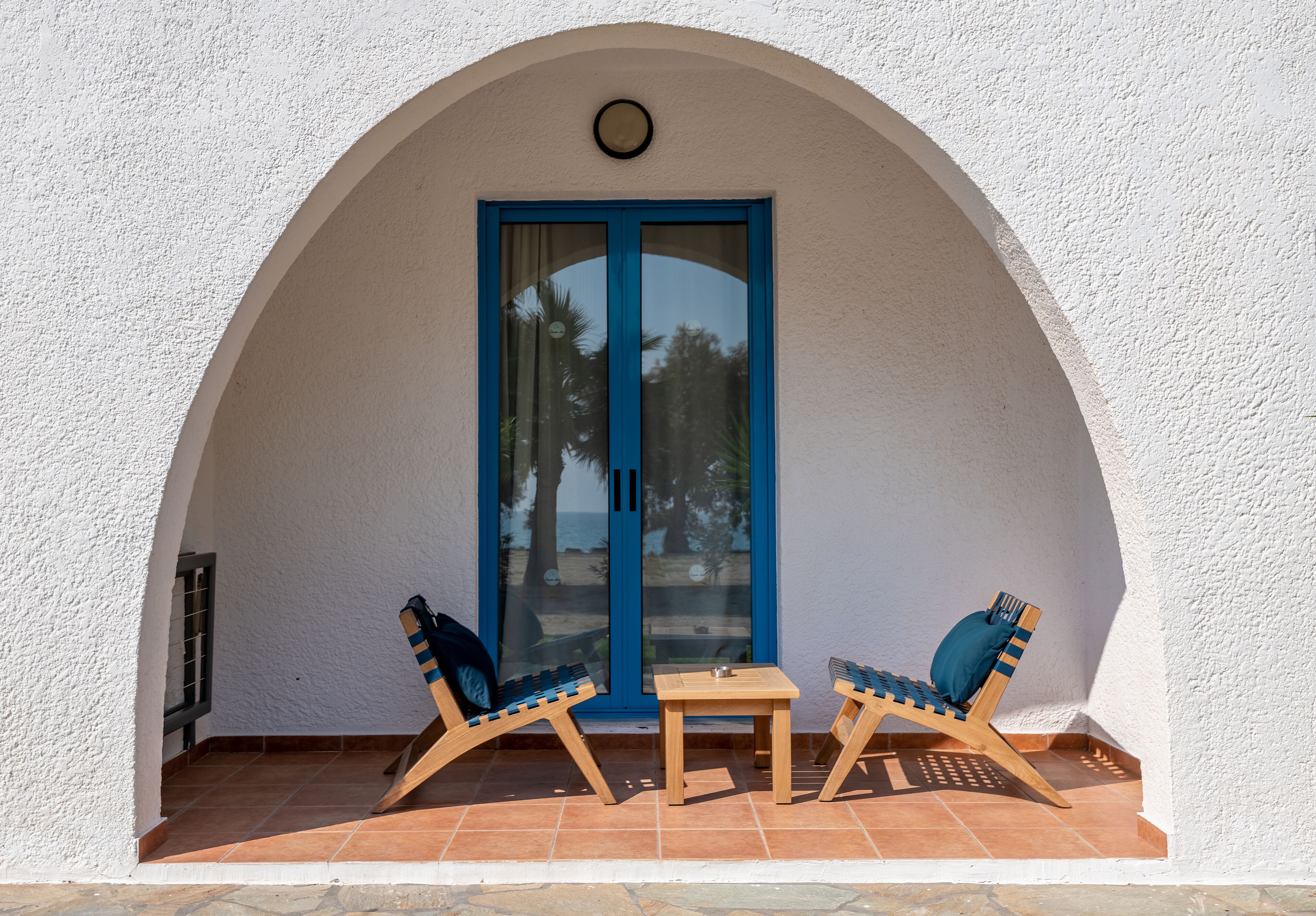 A small patio with two wooden chairs and a table, a blue-framed door, and a white textured wall.