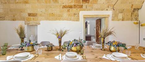 Una mesa de comedor dispuesta para seis personas, con platos, copas y cubiertos dorados y blancos. Hay dos jarrones con plantas y flores secas. La habitación tiene una pared de ladrillo y un techo de madera.