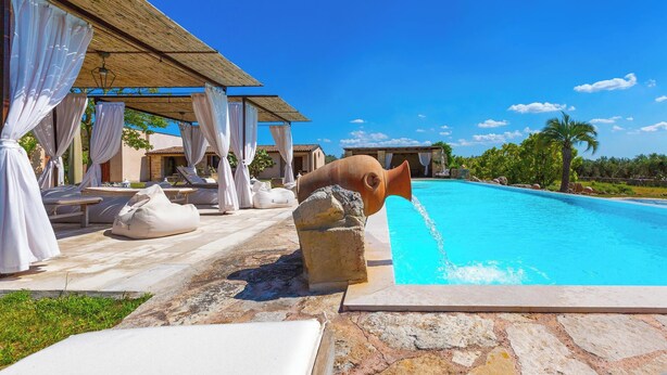 A poolside area with lounge chairs, a stone wall, and a building with a thatched roof. - 2874 Masseria Marchese by Perle di Puglia (Montesano Salentino)