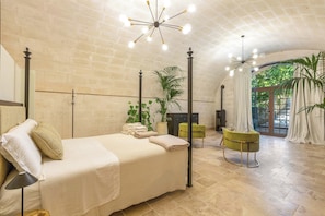 A spacious bedroom with a large bed, a chandelier, and a view of greenery through an arched doorway.