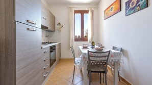 A kitchen with a dining area, featuring a table with chairs, a window with curtains, and a refrigerator. - 2989 Villetta Mito - Mito A by Barbarhouse (Tricase)