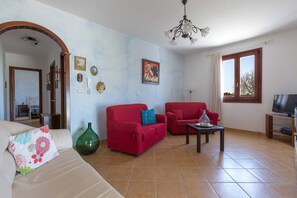 Uma sala de estar com um sofá, duas poltronas vermelhas, um vaso verde e uma TV sobre um suporte de madeira.