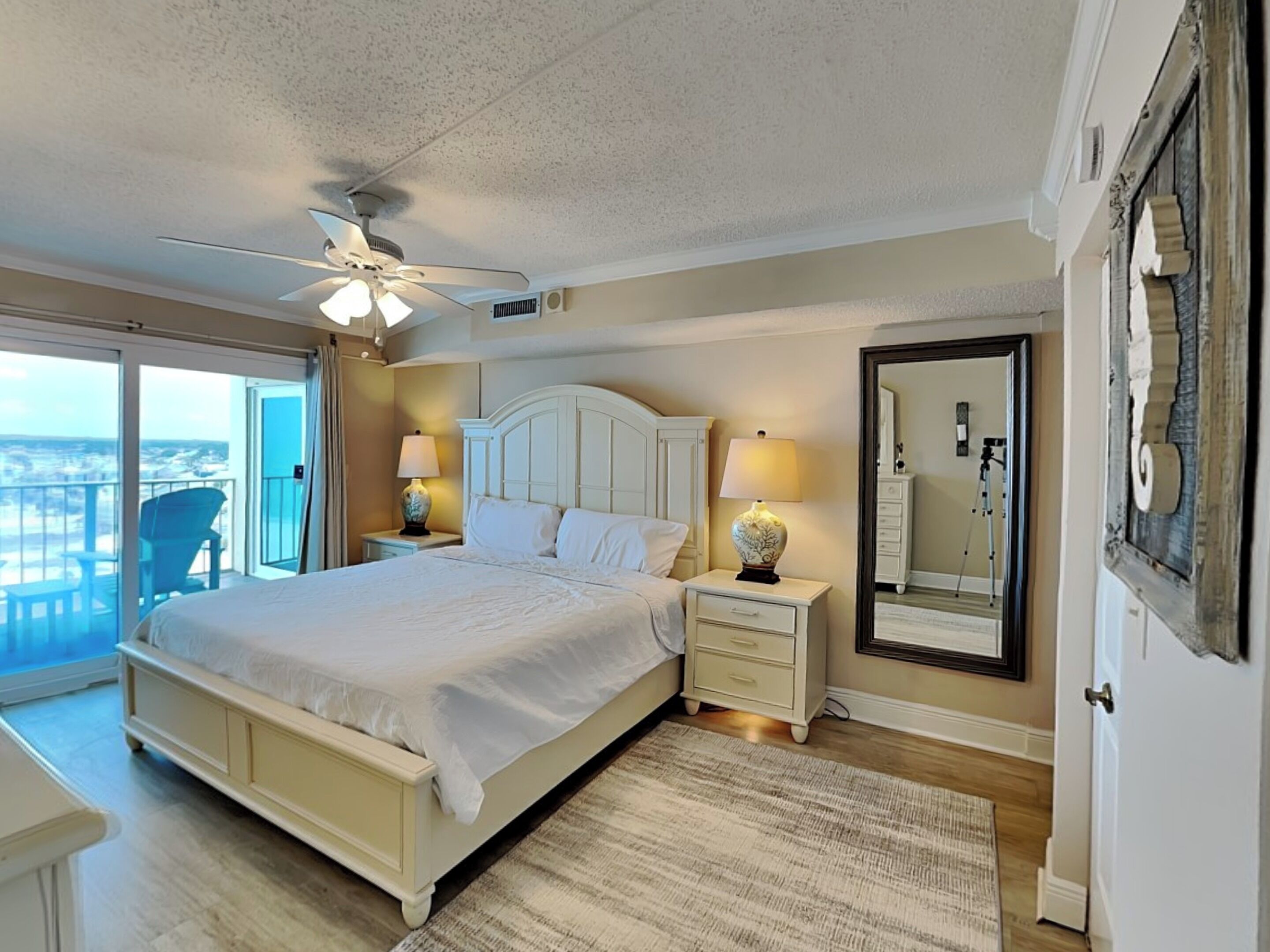 A bedroom with a large bed, two bedside tables with lamps, a ceiling fan, a balcony with a chair, and a large mirror on the wall.