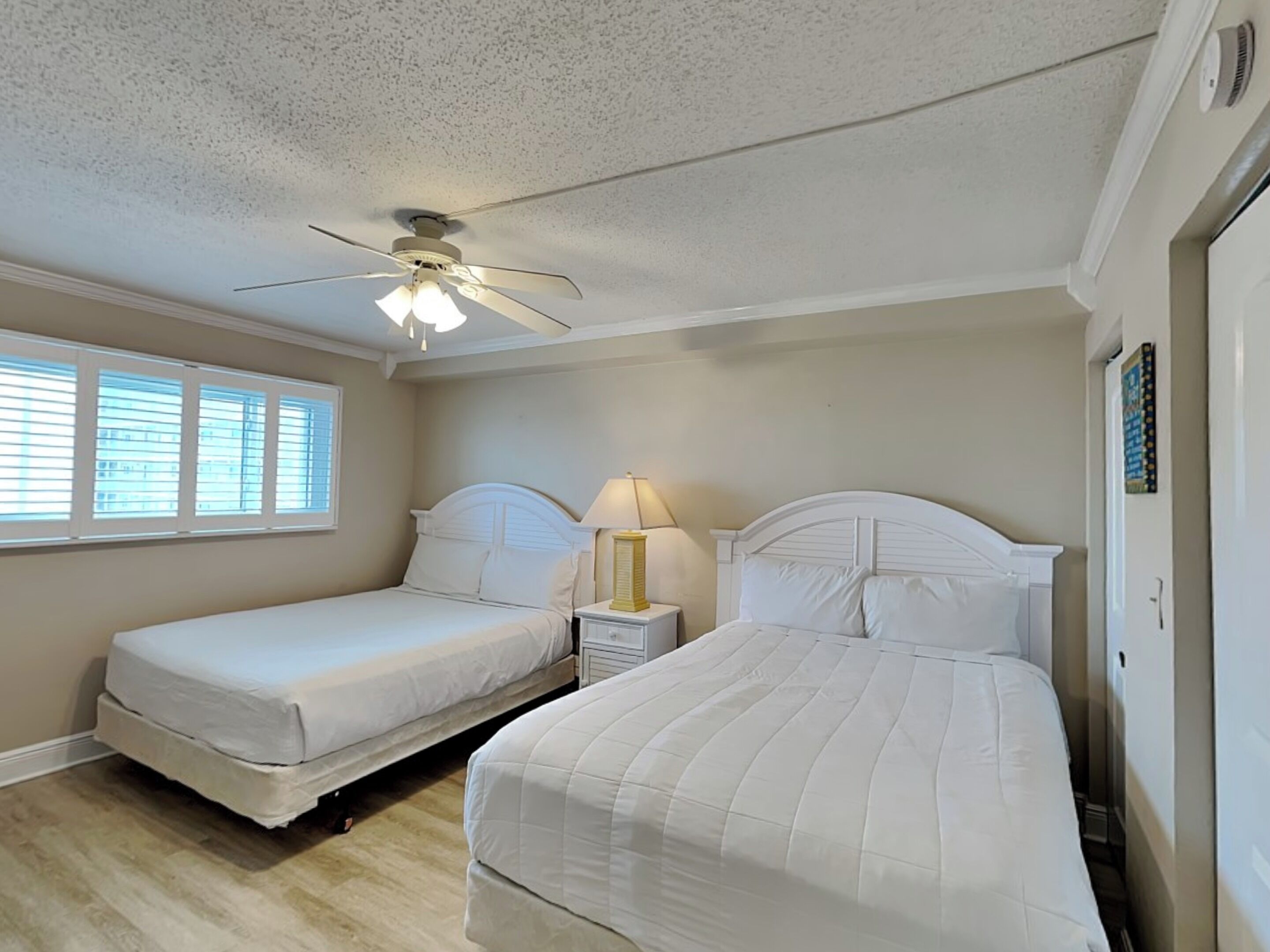 A bedroom with two beds, a ceiling fan, a nightstand with a lamp, and a window with blinds.