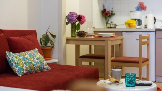 A cozy room with a red sofa, a small table with a cup and a remote control, and a kitchen area with a dining table and chairs. - Sweetest Thing Apartment (Dubrovnik)