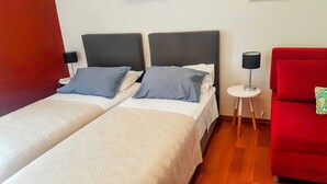 A hotel room with two single beds, a red armchair, a small round table with a lamp, and a red wall. - Sweetest Thing Apartment (Dubrovnik)