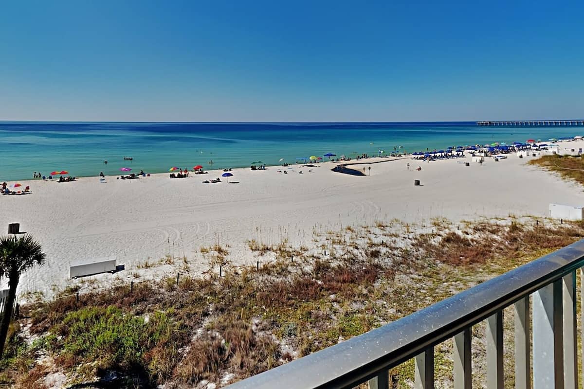 A beach with a pier in the distance, sandy beach, and some vegetation in the foreground.
