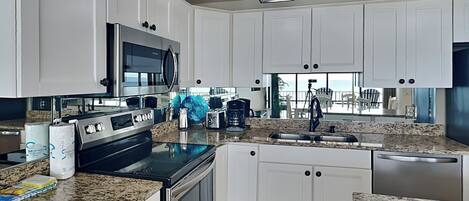 A modern kitchen with white cabinets, granite countertops, stainless steel appliances, and a large window offering a view of the outdoors.