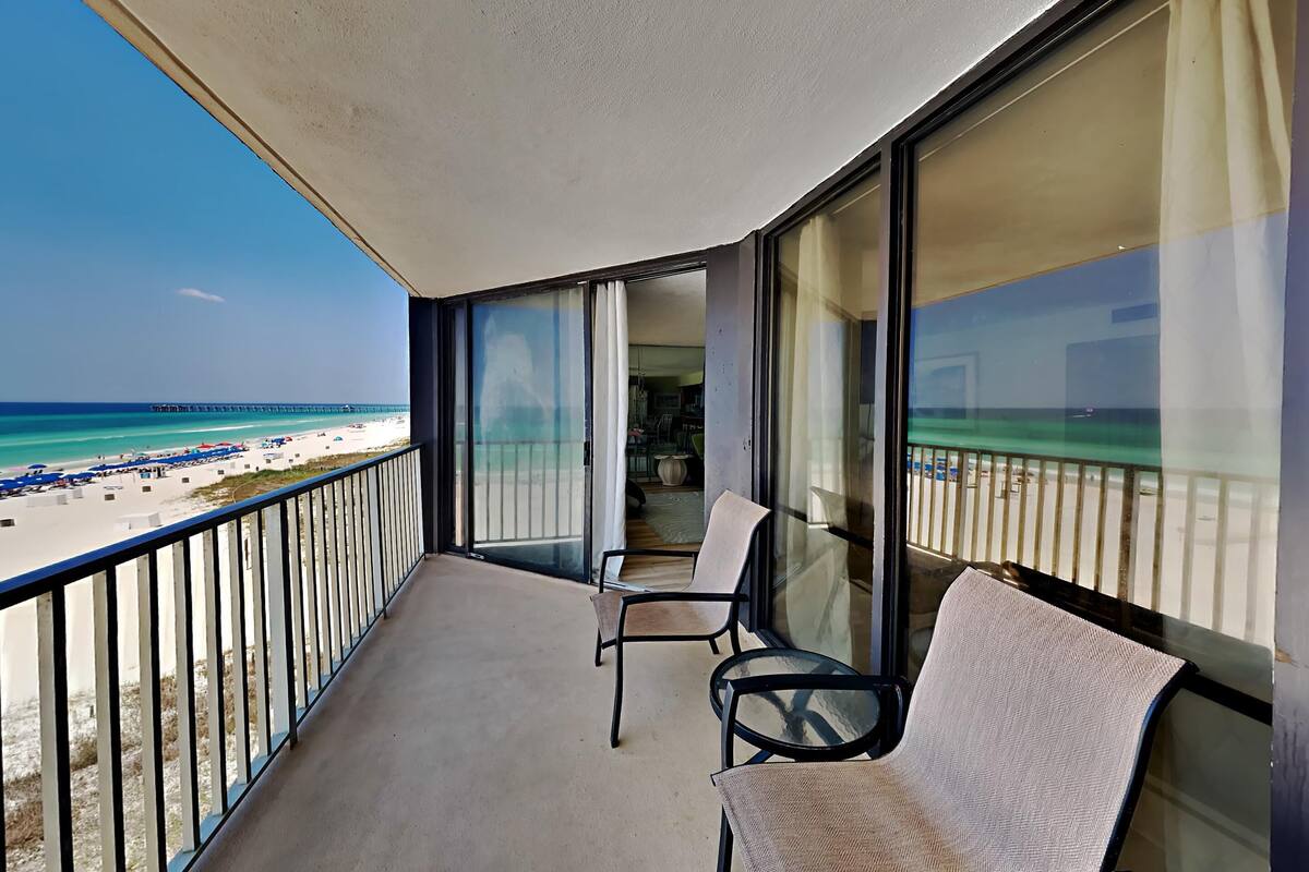 A balcony with a view of a beach and pier. There are two chairs and a table.