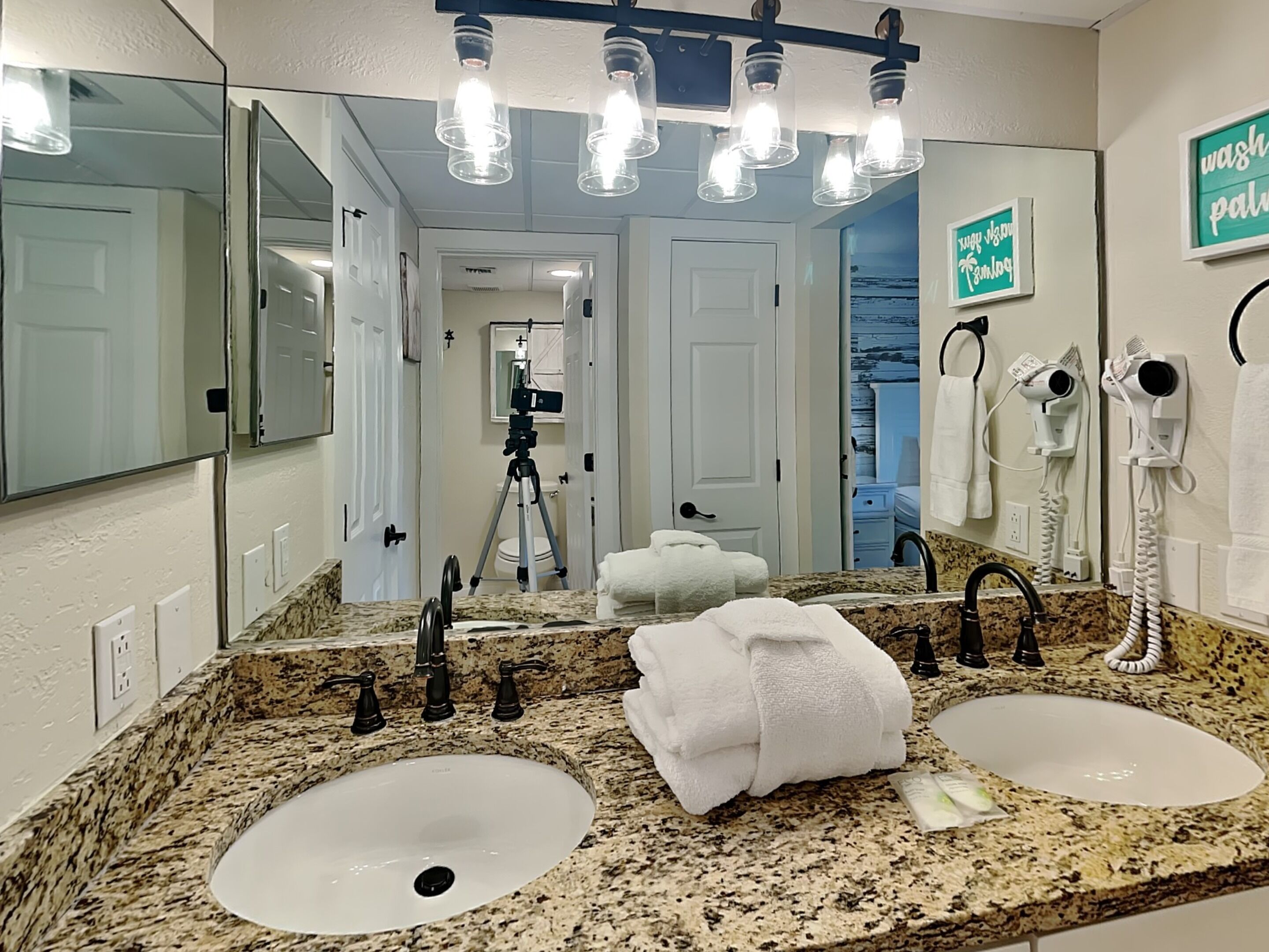 A bathroom with two sinks, a large mirror, and a vanity with a granite countertop. There are towels on the counter and a hairdryer mounted on the wall.