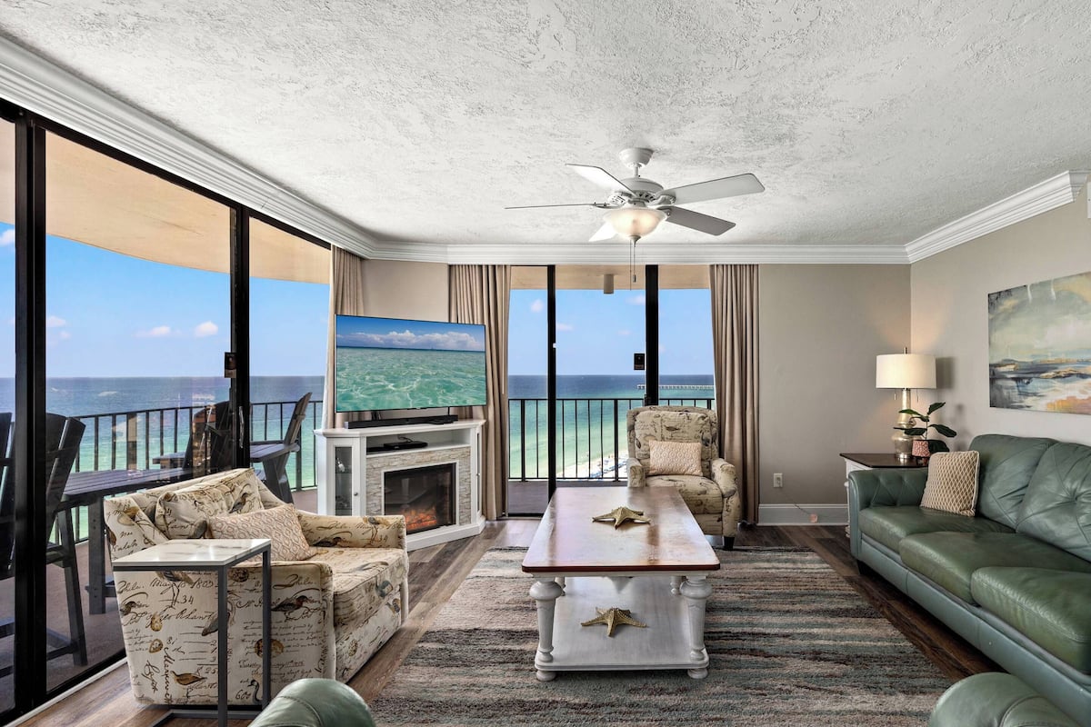 A spacious living room with a large TV, comfortable seating, a fireplace, and a balcony with a view of the ocean.