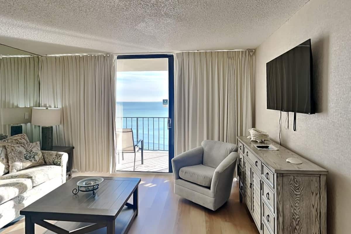 A hotel room with a balcony, a sofa, a chair, a TV, and a wooden cabinet.