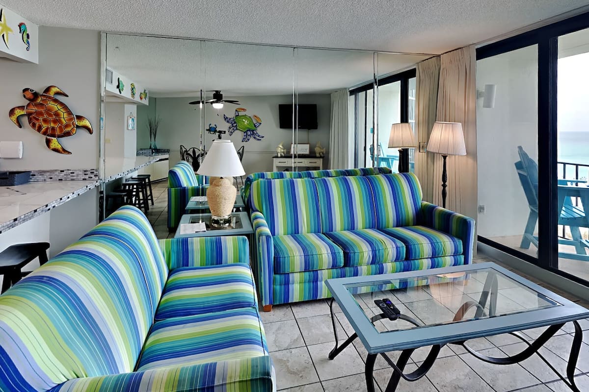 A living room with striped sofas, a glass coffee table, and a view of the ocean through large windows.