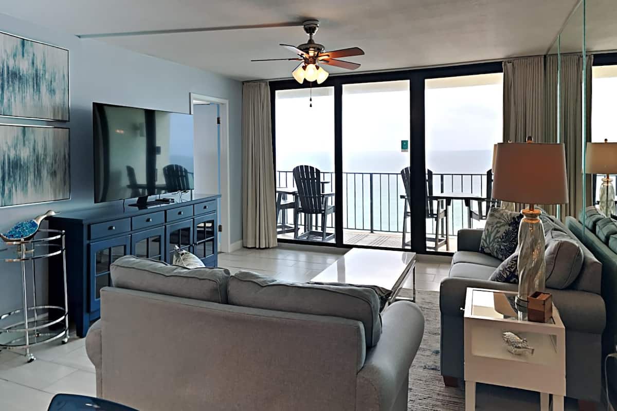 A modern living room with a large flat-screen TV, a ceiling fan, and a balcony with a view of the sea.