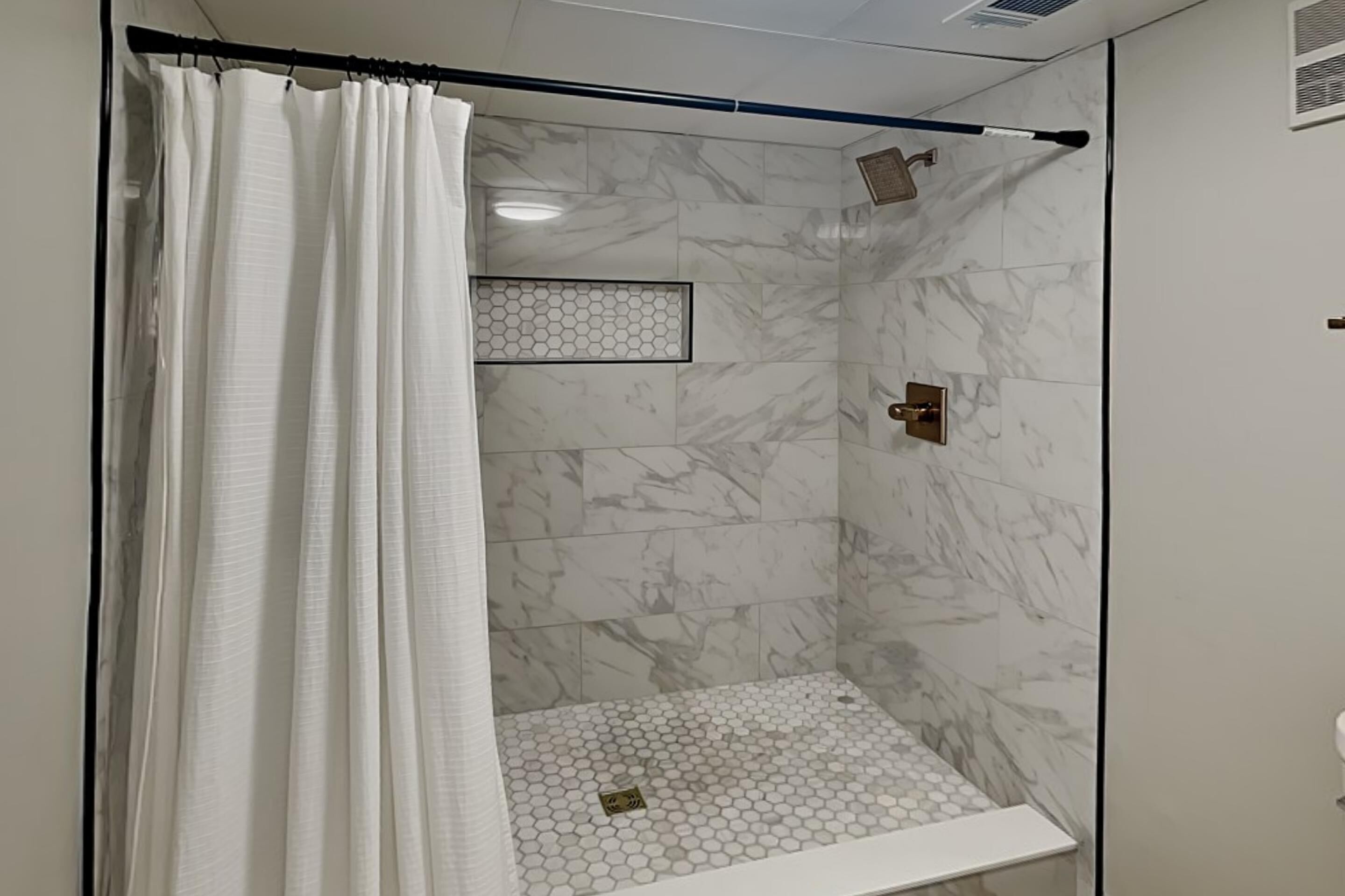 Una ducha con cortina blanca, suelo de azulejos hexagonales y paredes de mármol.