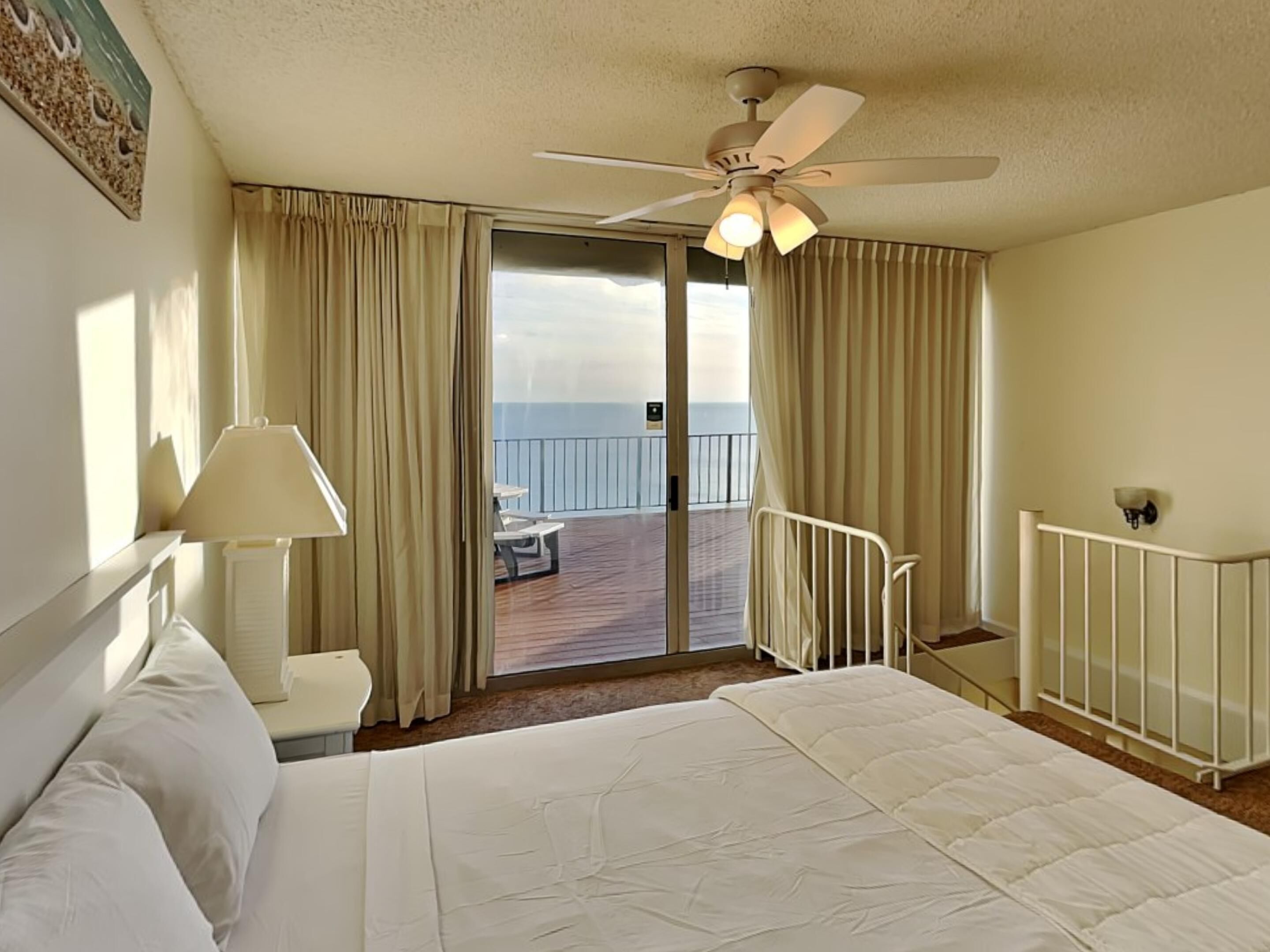 A hotel room with a bed, a desk, a chair, a lamp, a ceiling fan, and a balcony with a view of the ocean.