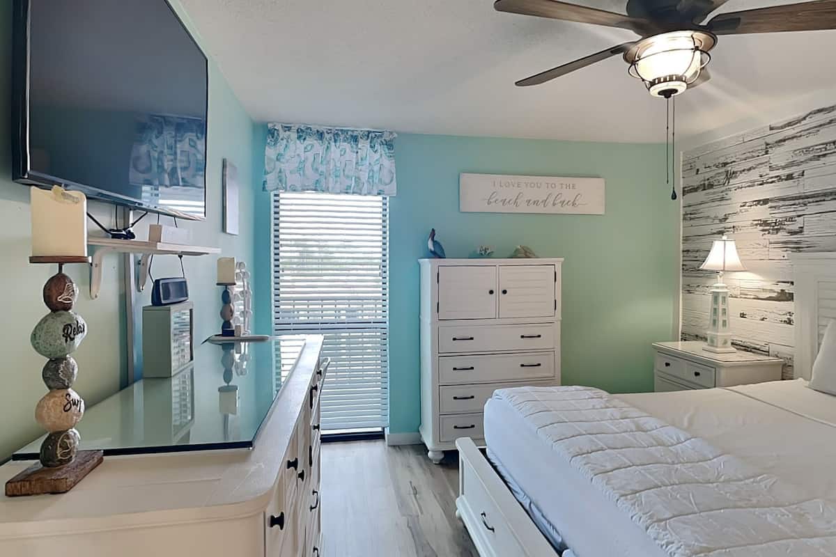 A bedroom with a bed, a dresser, a nightstand, a ceiling fan, a TV, and a window with blinds.
