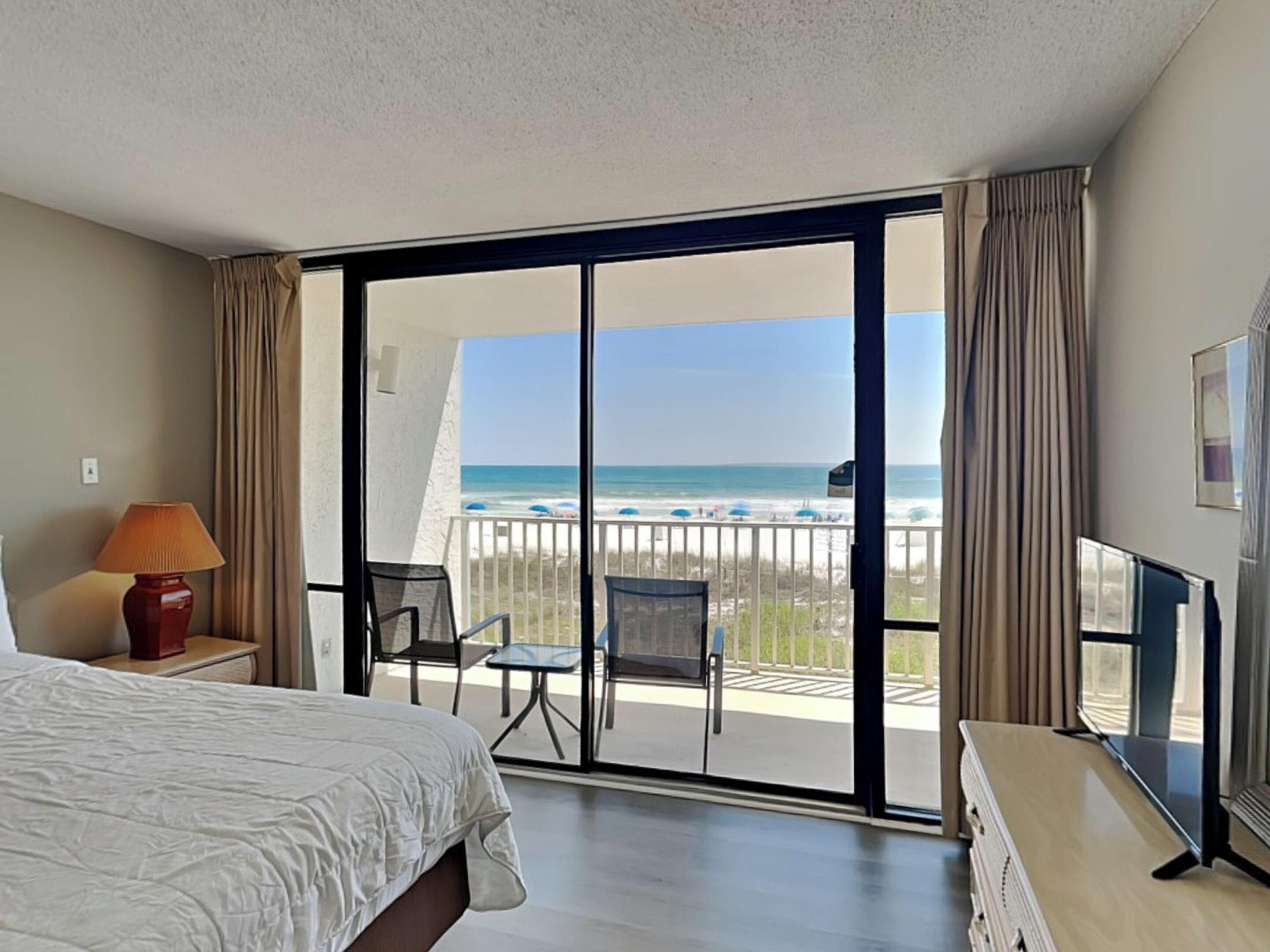 A hotel room with a bed, a desk with a lamp, a TV, and a balcony with a view of the beach and ocean.