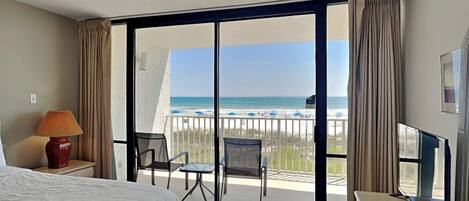 A hotel room with a bed, a desk with a lamp, a TV, and a balcony with a view of the beach and ocean.