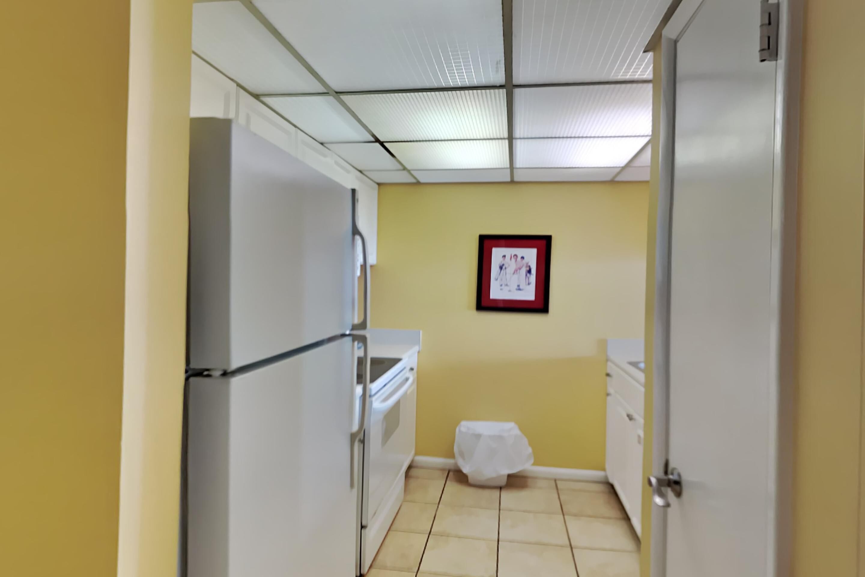 A small kitchen with a refrigerator, oven, and sink. There is a framed picture on the wall and a trash bin in the corner.