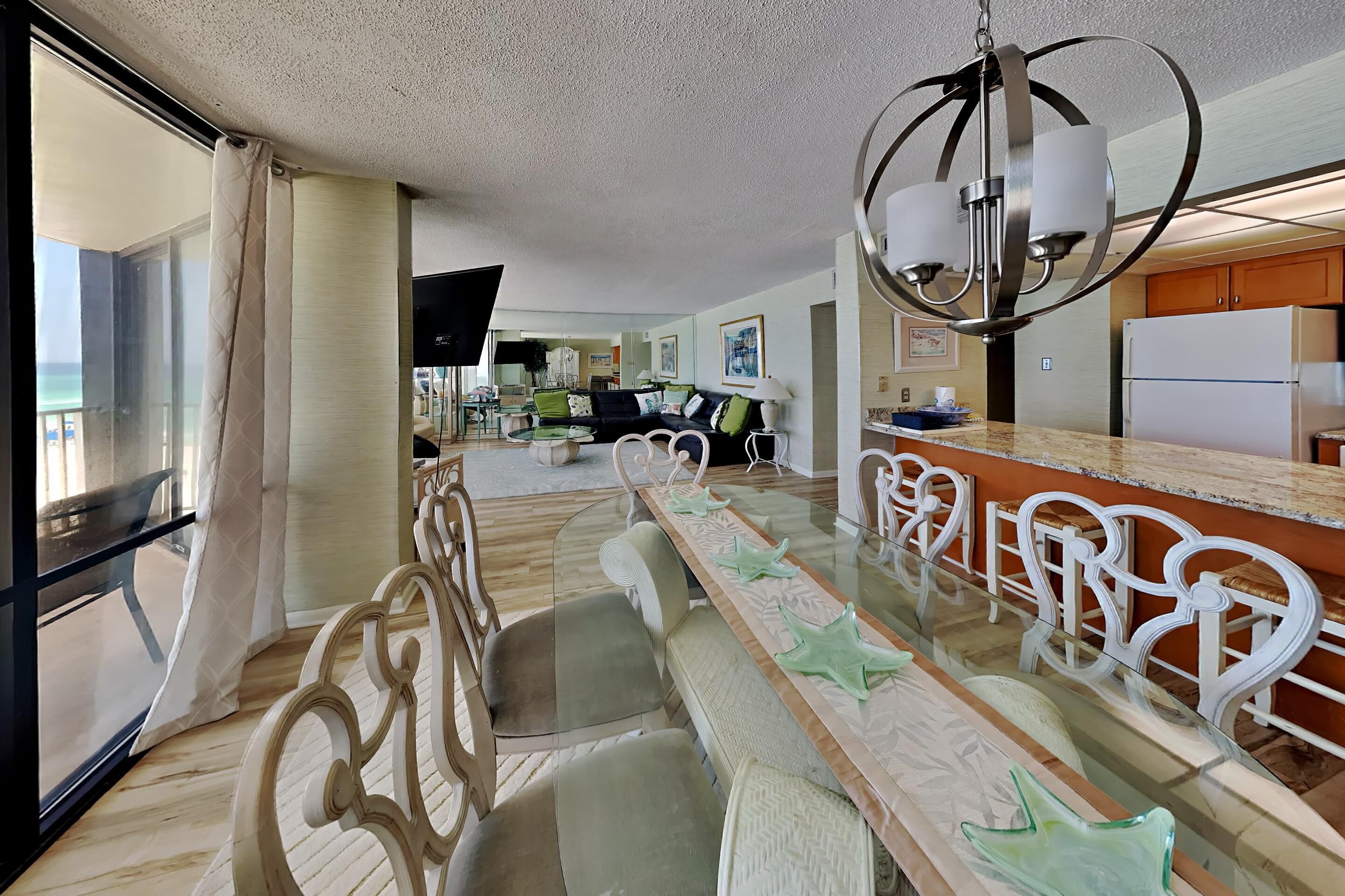 A spacious living room with a large glass dining table, comfortable seating, a kitchen area, and a balcony with a view.