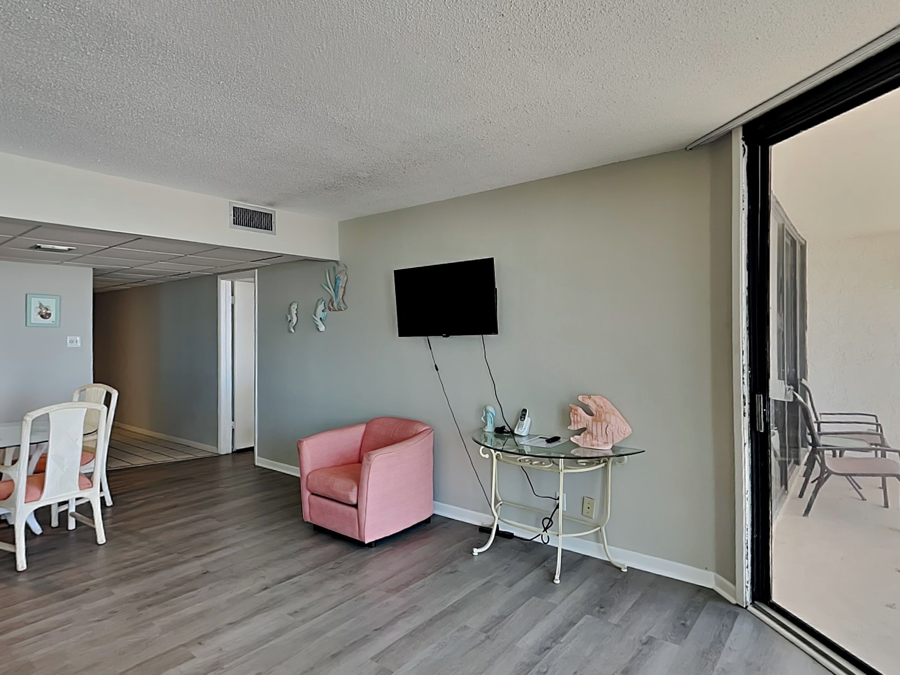 A modern living room with a pink chair, a glass table with a phone and decorative items, a flat-screen TV mounted on the wall, and a sliding glass door leading to an outdoor area.