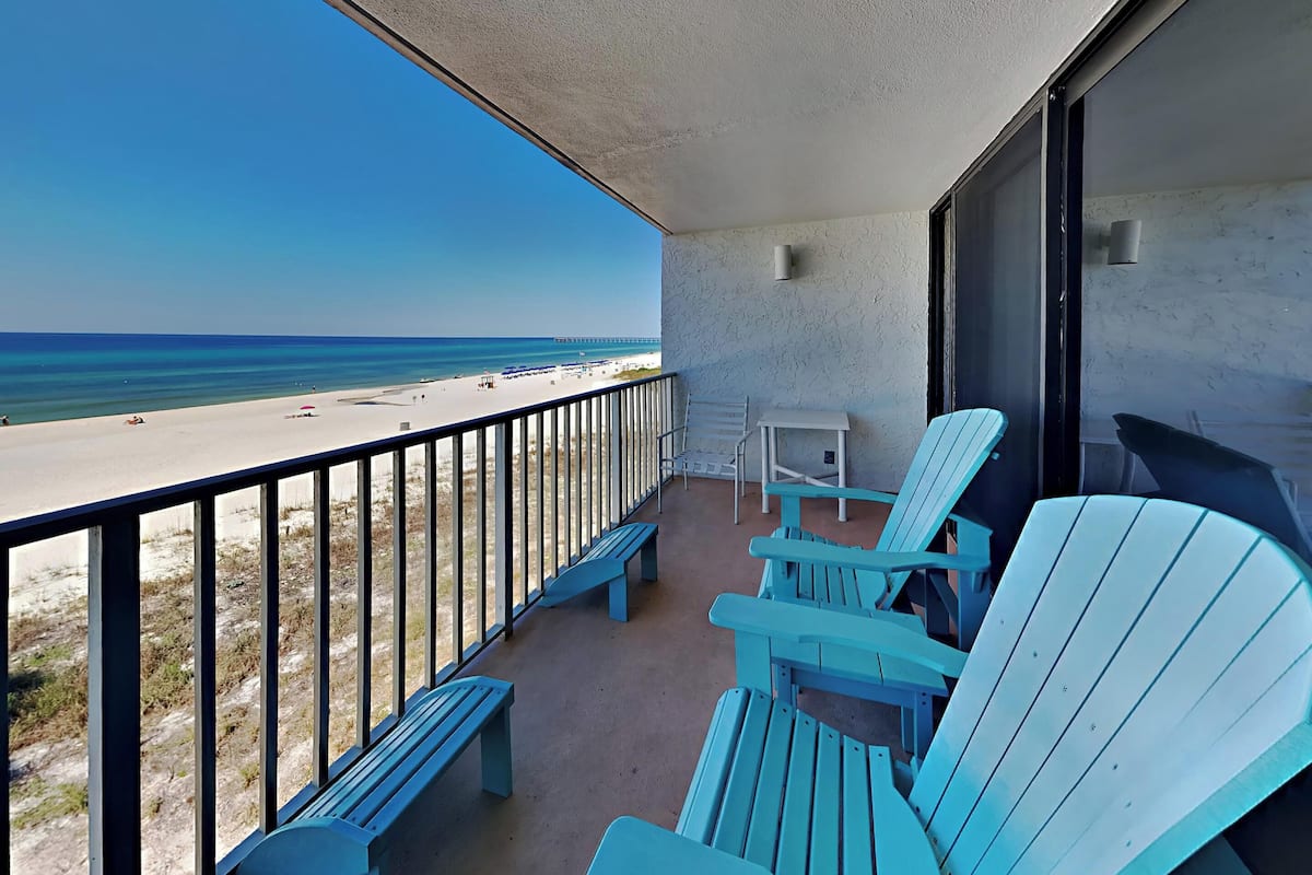 A balcony with a view of the beach, featuring blue Adirondack chairs and a small table.