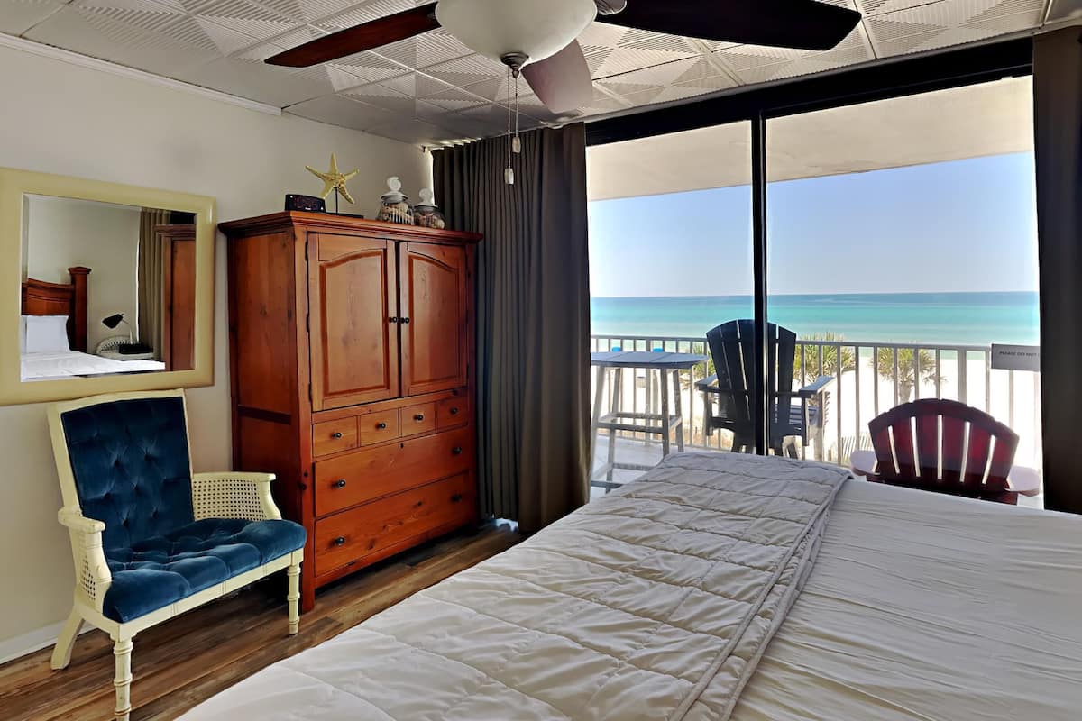 A bedroom with a bed, a wooden wardrobe, a blue chair, a large mirror, and a balcony with a view of the ocean.