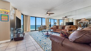 A spacious living room with a large window overlooking the ocean, a ceiling fan, and a television. - Edgewater Beach & Golf Resort VIII by Resort Collection (Panama City Beach)
