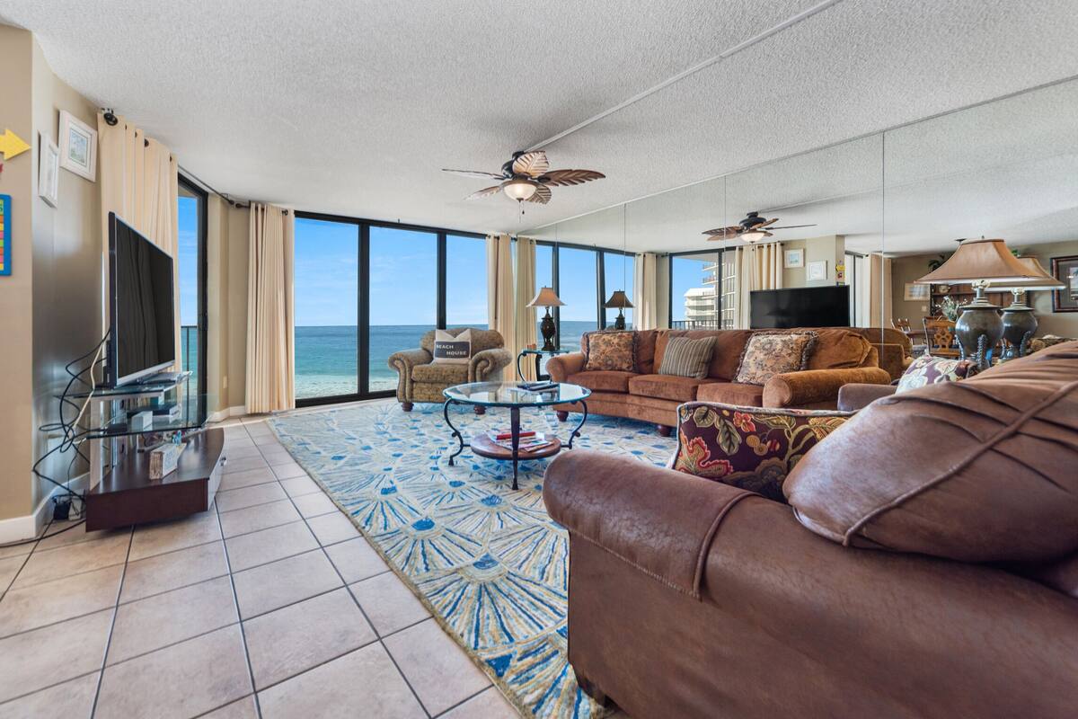 A spacious living room with a large window overlooking the ocean, a ceiling fan, and a television.