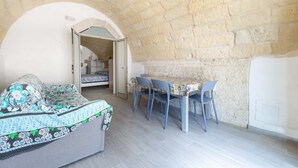 A room with a stone wall, a bed with a floral bedspread, a dining table with chairs, and a doorway leading to another room. - 2923 La Casa Della Nonna by Barbarhouse (Carpignano Salentino)