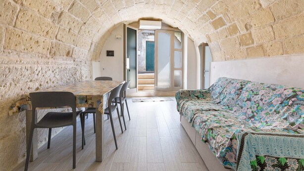 A cozy room with a stone arch ceiling, a dining table with chairs, a sofa with a floral blanket, and a view through an open door. - 2923 La Casa Della Nonna by Barbarhouse (Carpignano Salentino)